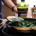 A bottle of oil is next to a pan on a portable burner that has stir-fried kale.