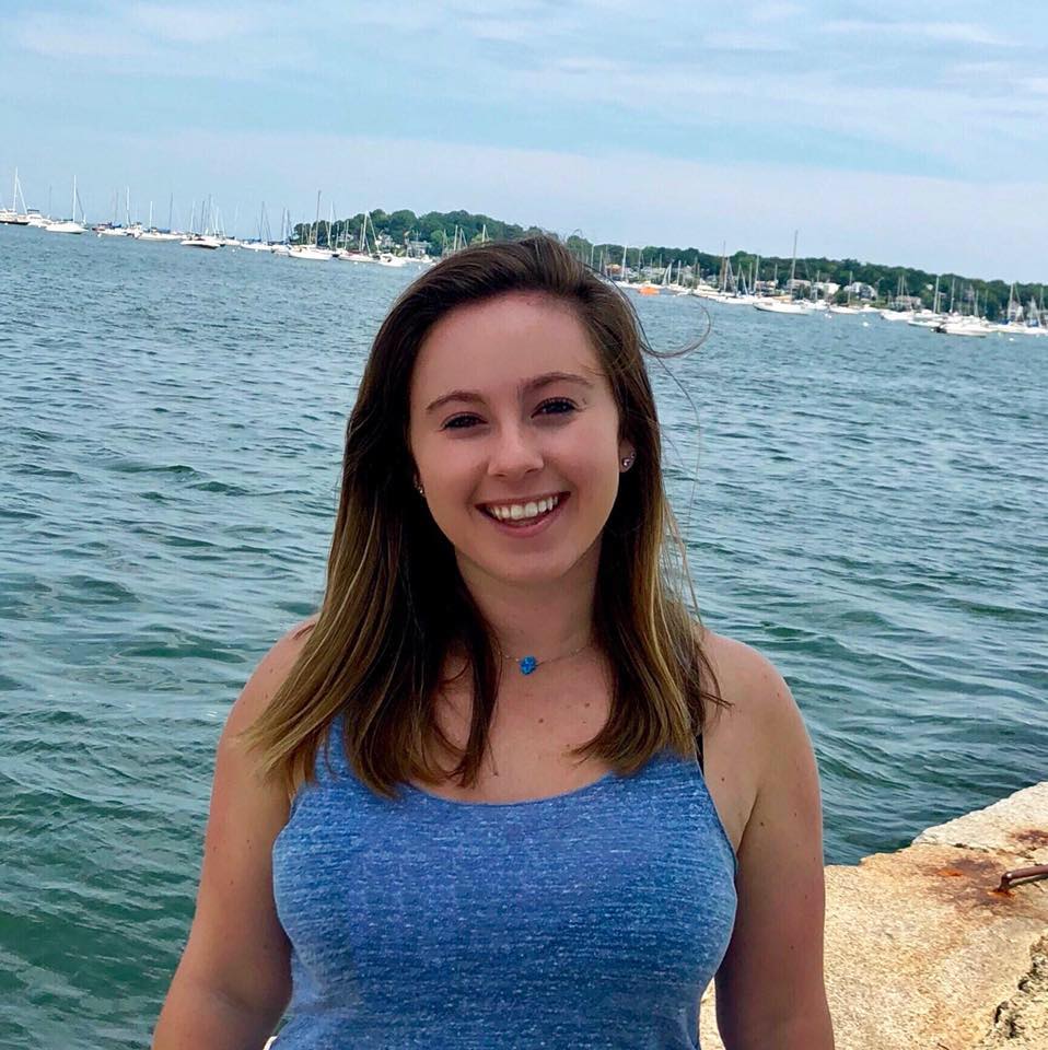 Ali is wearing a gray-blue tank top and is standing by a stone wall with the water of the Atlantic in the background.