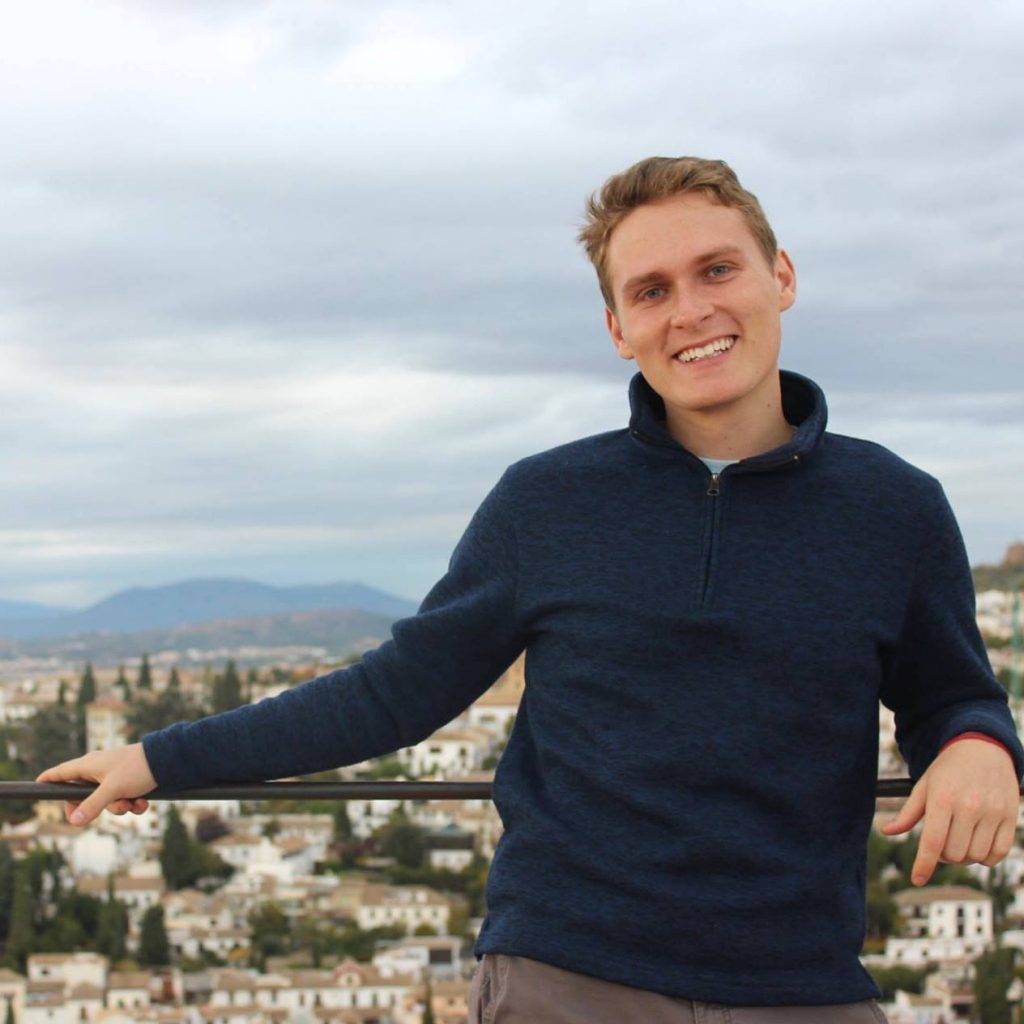 Kevin stands next to a railing that over looks a town. He is a wearing a navy blue sweatshirt.