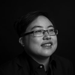 [Photo: Black and white image of a young East Asian person with glasses smiling and laughing, looking slightly away from the camera. Photo by Colin Pieters.]