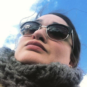low angle close-up of Amanda Lazarus with sunglasses & scarf, blue sky above